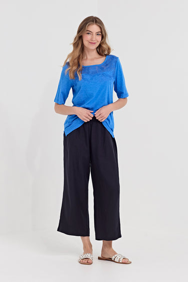 A woman modeling a bright blue short-sleeve top and dark navy wide-leg pants  by Verge against a light gray studio background.