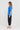 A woman stands in profile wearing a bright blue short-sleeve top and dark navy cropped pants by Verge with white slide sandals, photographed against a clean light gray studio background.