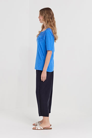 A woman stands in profile wearing a bright blue short-sleeve top and dark navy cropped pants by Verge with white slide sandals, photographed against a clean light gray studio background.