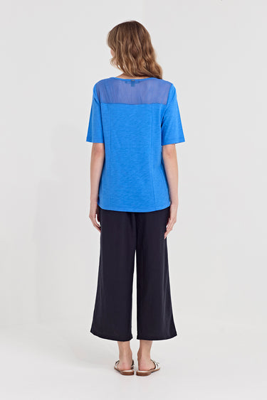 A woman viewed from the back wearing a bright blue short-sleeve top with a sheer yoke and dark wide-leg cropped pants by Verge, standing against a plain light gray studio background.