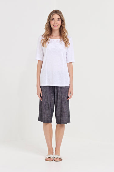 A woman modeling a white short-sleeve top and dark gray plaid cropped shorts by Verge against a plain light gray studio background.