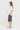 A woman standing in profile wearing a white short-sleeve top, dark gray plaid knee-length shorts by Verge, and white slide sandals against a plain white studio background.