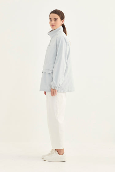 A woman stands in profile wearing a light icy blue lightweight jacket, white trousers by Verge, and white sneakers against a plain white studio background.