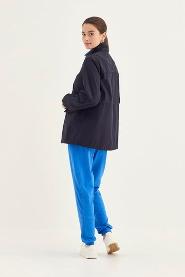 A woman modeling a dark navy blue lightweight jacket, paired with bright blue sweatpants by Verge and white sneakers, photographed from the back against a plain white studio background.