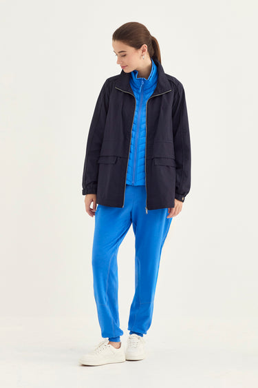 A woman modeling a dark navy zip-up jacket over a bright blue zip-up top, paired with matching blue sweatpants by Verge and white sneakers, against a plain white studio background.
