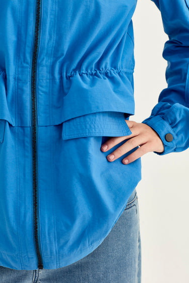 Close-up of a person wearing a blue lightweight jacket by Verge, showing the front zipper, gathered elastic detail, flap pocket, and hand tucked into the pocket.