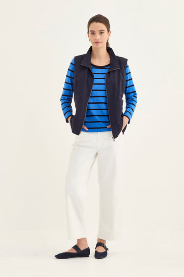 A woman modeling a navy blue sleeveless zip vest over a blue and navy striped long-sleeve top, paired with white wide-leg trousers by Verge and black flats, against a plain white studio background.