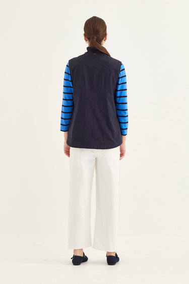 A woman viewed from the back wearing a sleeveless navy vest over a blue and navy striped long-sleeve top, paired with white wide-leg pants by Verge and dark flats, against a plain light background.