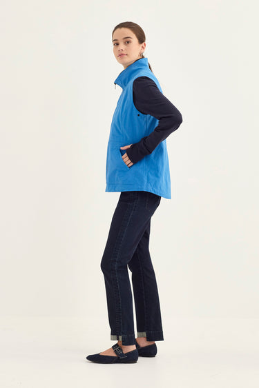 A woman modeling a bright blue sleeveless zip-front vest over a black long-sleeve top, paired with dark blue jeans by Verge and black flats, against a plain light background.