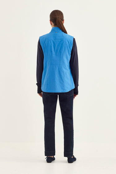A woman viewed from the back wearing a bright blue sleeveless vest over a dark navy long-sleeve shirt and dark navy pants by Verge, standing against a plain light background.