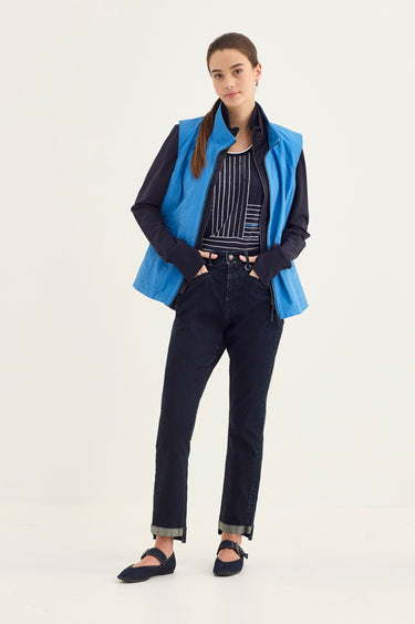 A model stands against a plain light background wearing a bright blue sleeveless vest over a black-and-white striped top, dark navy jeans by Verge, and dark flat shoes.