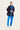 A woman modeling a dark navy blue lightweight jacket over a bright blue zip-up shirt, paired with bright blue joggers by Verge and white sneakers, against a plain white studio background.