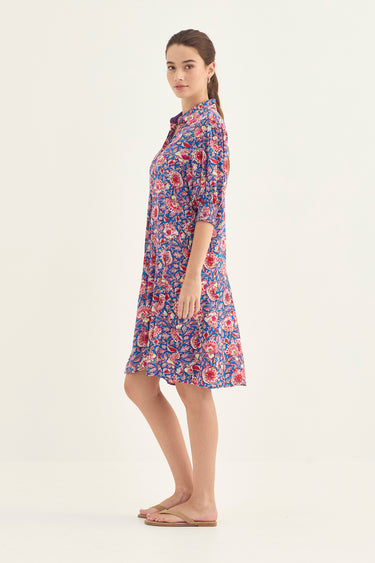 A woman modeling a short-sleeved button-up shirt dress by Verge with a dense red, pink, and white floral print on a blue-purple background, set against a plain light beige studio backdrop.