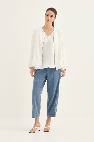 A woman modeling a white embroidered jacket over a white top, paired with light blue cropped pants by Verge and white heeled sandals, against a plain white studio background.