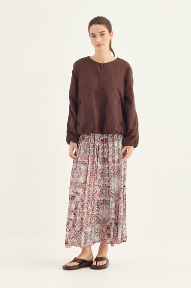 A model stands against a plain off-white studio background wearing a dark brown cropped long-sleeve top and a long pleated maxi skirt with a dusty rose and beige paisley pattern by Verge, paired with dark brown slide sandals.
