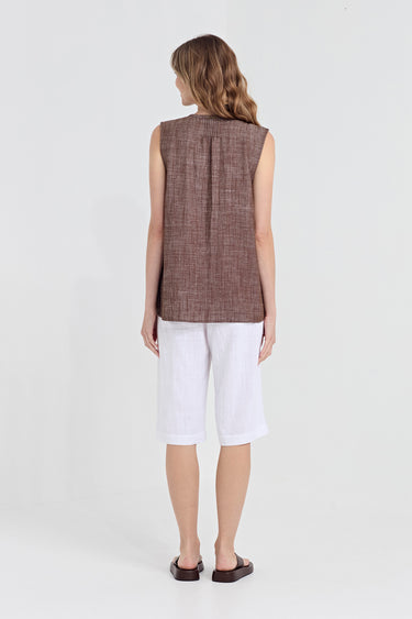 A woman viewed from the back wearing a sleeveless brown textured linen vest, white linen shorts by Verge, and dark brown sandals, set against a plain light gray studio background.