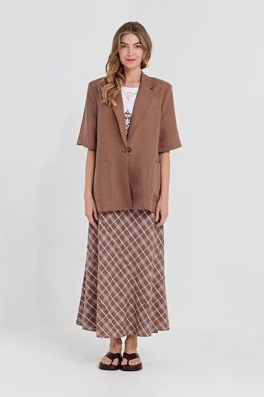 A woman modeling a brown short-sleeve blazer over a white patterned top and a brown plaid maxi skirt by Verge, standing against a light gray studio background.