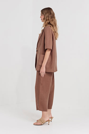 A woman stands in profile wearing a matching taupe-brown short-sleeve blazer and cropped wide-leg trousers set by Verge, paired with beige heeled sandals, against a plain light gray studio background.