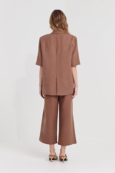 A woman viewed from the back wearing a matching taupe-brown short-sleeve blazer and wide-leg cropped pants set by Verge, paired with black and beige slingback heels, against a plain white studio background.
