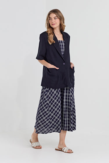 A woman modeling a dark navy blue short-sleeve blazer over a matching navy and white plaid dress by Verge, paired with white slide sandals, against a light gray studio background.