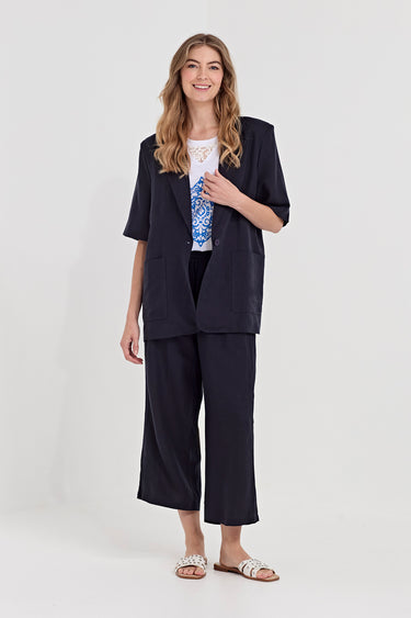 A woman modeling a dark navy blue two-piece suit consisting of a short-sleeve blazer and matching cropped trousers, styled with a white t-shirt and patterned blouse by Verge, against a plain light gray studio background.