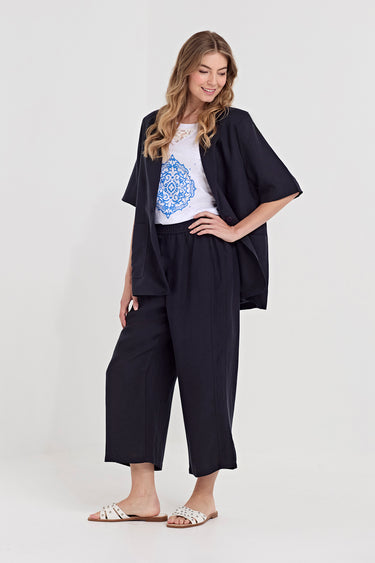 A woman modeling a black two-piece lounge set consisting of a short-sleeve open-front cardigan and matching cropped pants, styled over a white graphic t-shirt by Verge and white slide sandals, against a light gray studio background.