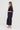 A woman stands in profile wearing a dark navy two-piece outfit consisting of a short-sleeve button-front jacket and matching wide-leg cropped pants by Verge, paired with white slide sandals, against a plain light gray studio background.