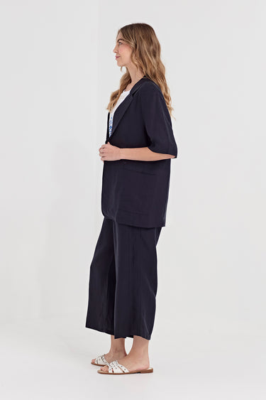 A woman stands in profile wearing a dark navy two-piece outfit consisting of a short-sleeve button-front jacket and matching wide-leg cropped pants by Verge, paired with white slide sandals, against a plain light gray studio background.