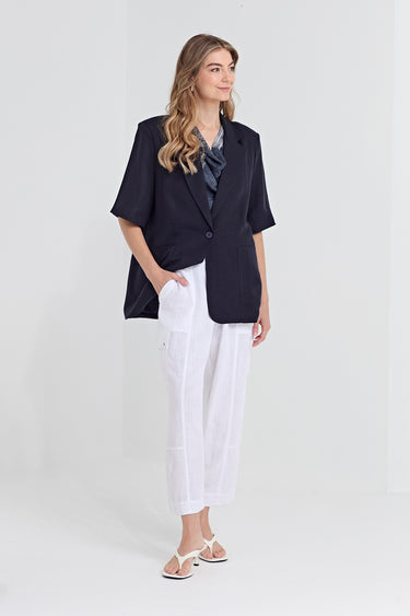 A woman modeling a short-sleeve navy blazer, white cropped pants, a blue patterned blouse by Verge, and white sandals against a plain white studio background.