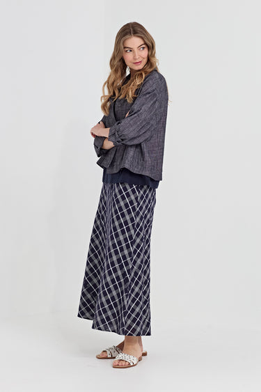 A woman modeling a two-piece outfit consisting of a dark gray textured long-sleeve top and a navy blue and white plaid maxi skirt by Verge, paired with white slide sandals, against a light gray studio background.