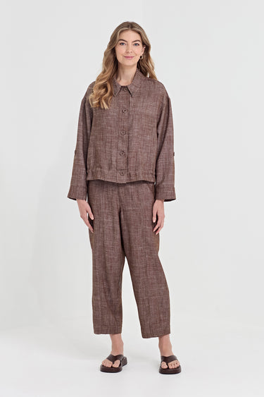 A woman modeling a matching brown textured two-piece set consisting of a cropped button-front jacket and cropped pants by Verge, paired with dark brown sandals, against a light gray studio background.