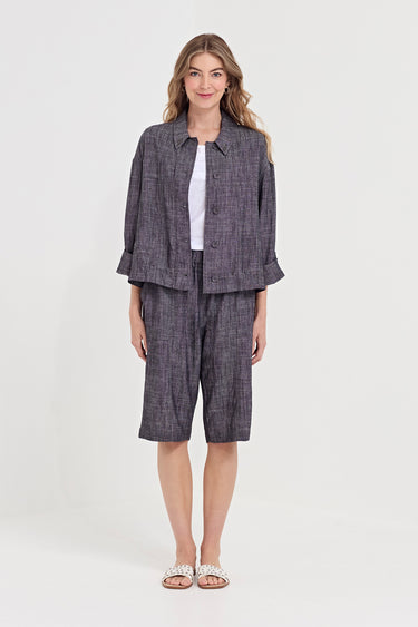 A woman stands against a light gray studio background wearing a matching two-piece outfit consisting of a cropped button-front jacket and shorts  by Vergin a dark gray and black textured plaid pattern, styled over a white top and white slide sandals.