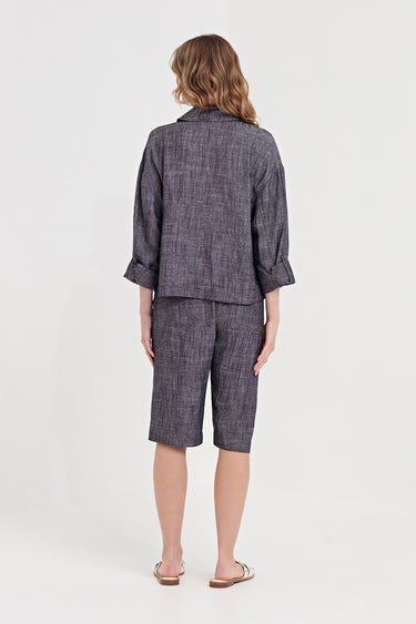 A woman viewed from the back wearing a matching dark gray and black textured linen-blend set consisting of a cropped jacket with rolled sleeves and matching cropped shorts by Verge.