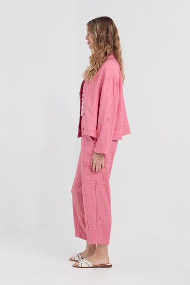 A woman stands in profile wearing a coordinated pink outfit consisting of a cropped button-front jacket and matching wide-leg cropped pants by Verge, paired with white slide sandals, against a plain light gray studio background.