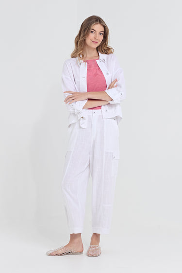 A woman modeling a white linen-blend two-piece set consisting of a cropped button-up shirt with a tie waist and matching relaxed-fit pants by Verge, worn over a pink top.