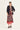 A woman modeling a long-sleeve midi dress by Verge with a colorful paisley and floral print in purple, orange, and brown tones, featuring a solid dusty rose gradient hem. She is styled with a black blazer and black slide sandals against a plain off-white studio background.