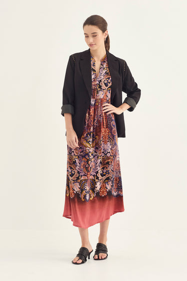 A woman modeling a long-sleeve midi dress by Verge with a colorful paisley and floral print in purple, orange, and brown tones, featuring a solid dusty rose gradient hem. She is styled with a black blazer and black slide sandals against a plain off-white studio background.