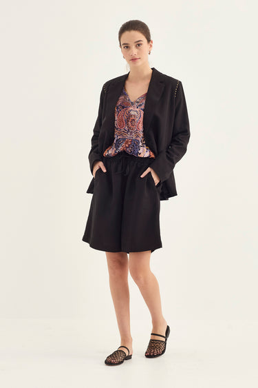 A model stands against a plain white studio background wearing a black tailored blazer and matching black shorts over a colorful paisley-patterned top by Verge, with black lace-up flat shoes.