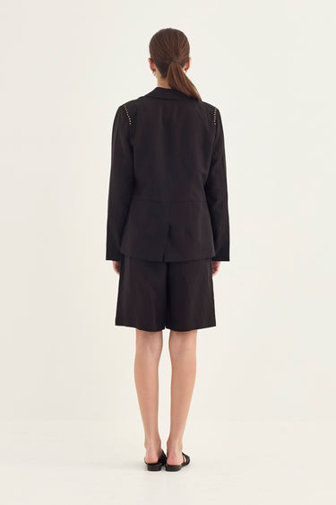 A model viewed from the back wearing a black tailored blazer and matching shorts set by Verge, with small gold chain details visible on the shoulders, against a plain off-white studio background.