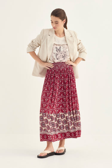 A woman modeling a long, pleated maxi skirt with a red and cream floral block print, styled with a white printed top, a beige blazer by Verge, and black slide sandals against a plain off-white studio background.