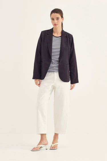 A woman modeling a dark navy blazer over a black and white striped top, paired with white cropped trousers by Verge and white slide sandals, against a plain light gray studio background.