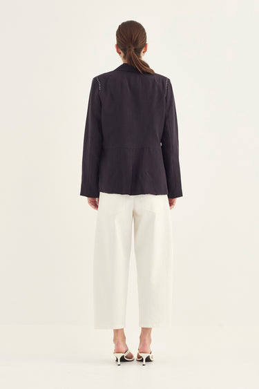 A model shown from the back wearing a dark charcoal gray blazer and white cropped trousers by Verge, standing against a plain off-white studio background.
