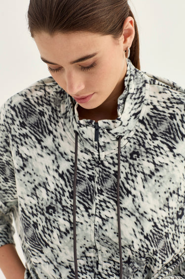 A close-up of a woman wearing a snake print jacket patterned by Verge with black drawstrings, looking down against a plain light background.