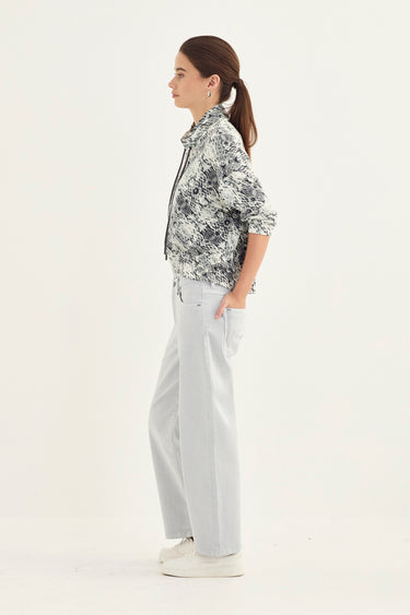 A woman stands in profile wearing a snake print jacket pattern with a button-up shirt with rolled sleeves, light gray wide-leg trousers by Verge, and white sneakers against a plain white studio background.