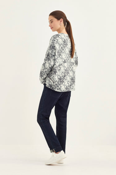 A woman is shown from a side-back angle wearing a snake print sweater and dark blue jeans by Verge, and white sneakers against a plain white studio background.