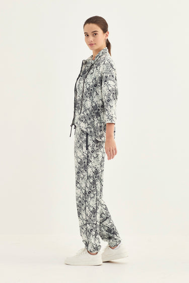 A woman modeling a matching two-piece set with a snake print jacket pattern, consisting of a long-sleeve zip-up hoodie and matching pants by Verge, paired with white sneakers against a plain white studio background.
