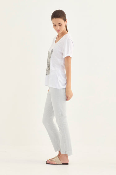 A woman modeling a white short-sleeve V-neck t-shirt with a subtle graphic print on the front, paired with light gray cropped pants by Verge and beige flat sandals, set against a plain white studio background.