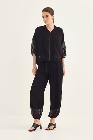 A model wearing a black two-piece outfit consisting of a cropped zip-up jacket with sheer mesh sleeves and matching loose black trousers with sheer mesh cuffs, paired with black slide sandals by Verge, against a plain off-white studio background.