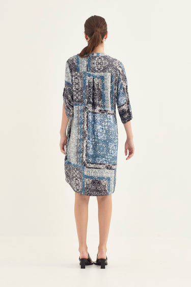 A woman viewed from the back wearing a knee-length patchwork print dress by Verge with three-quarter sleeves and black flat shoes, photographed against a plain light studio background.