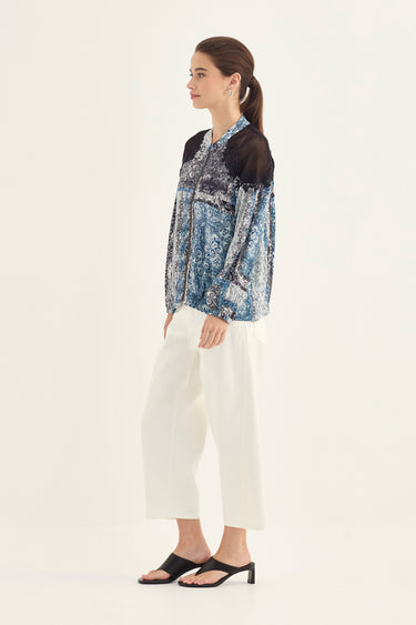 A woman stands in profile wearing a blue and white patterned long-sleeve blouse with black shoulder panels, white cropped pants by Verge , and black heeled sandals against a plain light background.